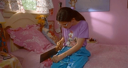 Dawn Wiener, a nerdy girl from Welcome To The Dollhouse, wearing glasses and t-shirt and jeans, sawing the head off a Barbie doll.