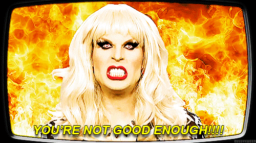 Blonde drag queen Katya surrounded by flames, with devil eyes, shouting "You're not good enough!!!!" before smiling warmly.