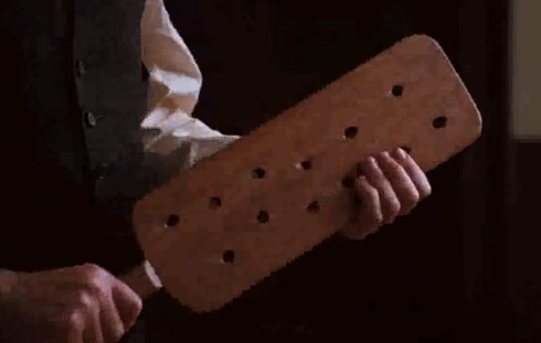 A smartly-dressed man holds a large wooden paddle with holes in it.