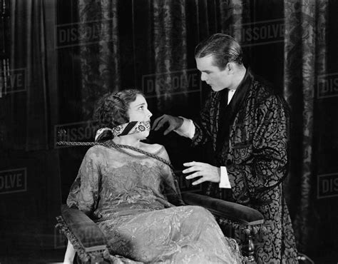 Vintage 1930s photo of a young woman with short hair, tied to a chair in front of a theatre curtain. She look anxiously at a smart young man in a silk dressing gown as he adjusts her cloth gag.