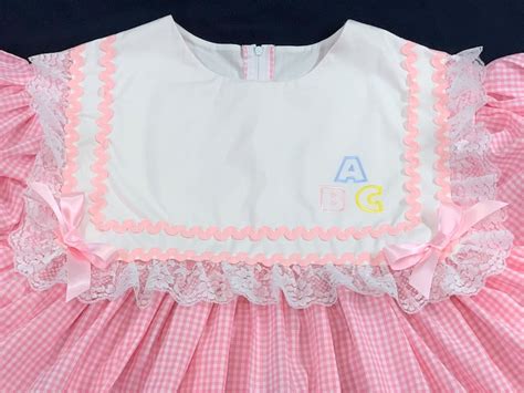 ABDL adult baby dress in pink gingham with a white frilly collar and ABC blocks embroidered on it.