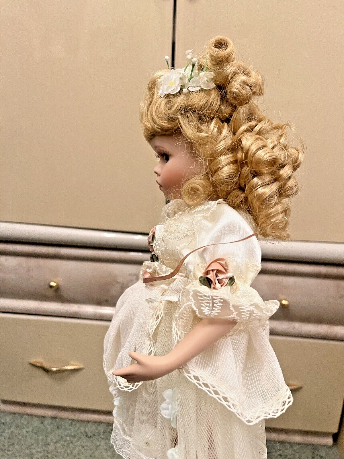 A vintage baby doll with curly blonde hair and a frilly white dress, standing side-on, head slightly down.